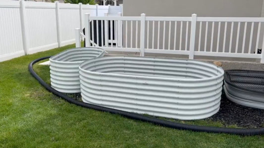 Two large, white corrugated metal tanks in a grassy area with a black hose running alongside them and a white fence in the background.